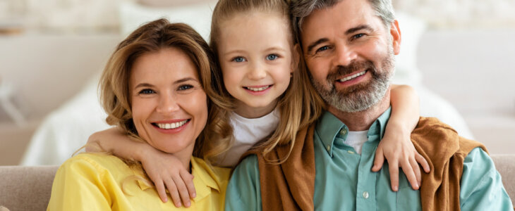 Shot Of Cheerful Family Of Three. Parents And Little Daughter Smiling To Camera Embracing Sitting On Couch At Home. Kid Hugging Her Dad And Mom Bonding On Weekend. Parenthood Concept