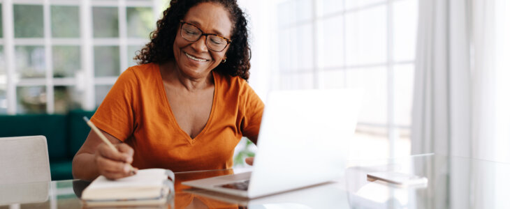Happy senior woman sitting at a table in her home office, drafting her last will and testament in a journal and taking care to allocate her assets. Retired woman leaving a plan in place for the future.